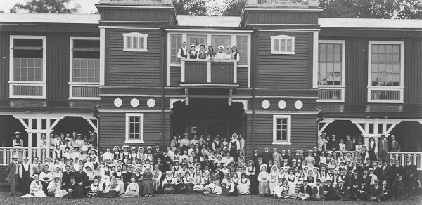 hundreds of men and women standing in front of large wooden building